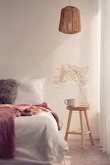 Bedside table with mug and flower next to bed with white bedding fur pillow and pink blanket, real photo with copy space on the empty wall