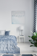 Poster above cabinet with lamp next to blue bed in white simple bedroom interior with plant. Real photo
