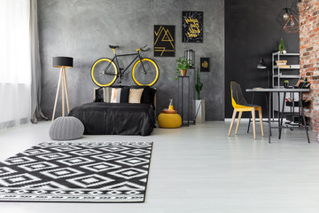 Patterned carpet and posters in grey bedroom interior with bike above black bed near lamp. Real...