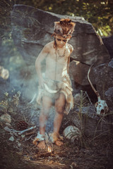 Caveman, manly boy at the fire. Scary young primitive boy outdoors near bonfire. Witch craft concept. Angry caveman, manly boy with horns near bonfire. Prehistoric tribal man outdoors on nature