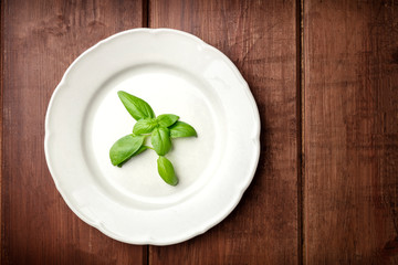 An overhead photo of a white plate with fresh basil leaves, shot from the top on a dark rustic wooden background with copy space