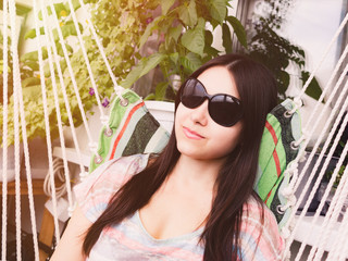Beautiful girl with long dark hair in sunglasses is resting sitting in a hammock on the balcony