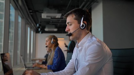 Attractive business man in headset calling to cuctomers in business office call center. His female colleagues also calling and communicated by phone with clients. 4k slow motion, - Powered by Adobe