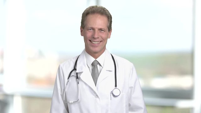 Smiling middle aged doctor portrait, front view. Meture man working as pshysician wearing medical suit and stethoscope. Abstract blurred background.