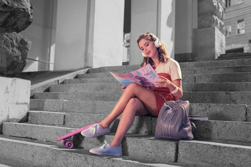 Fototapeta na wymiar Magazine and music. Modern fashionable girl sitting on the stairs outside reading fashion magazine and listening to music