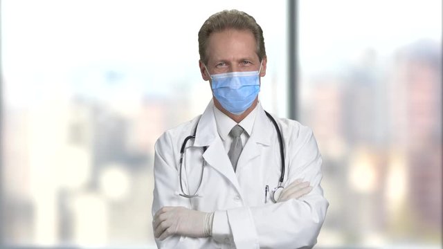 Portrait of physician in mask with folded arms. Doctor with stethoscope in white coat suit. Bright abstract blurred windows background with view on city.