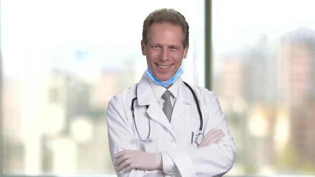 Mature middle aged smiling doctor folding arms. Bright abstract blurred windows background with view on city.