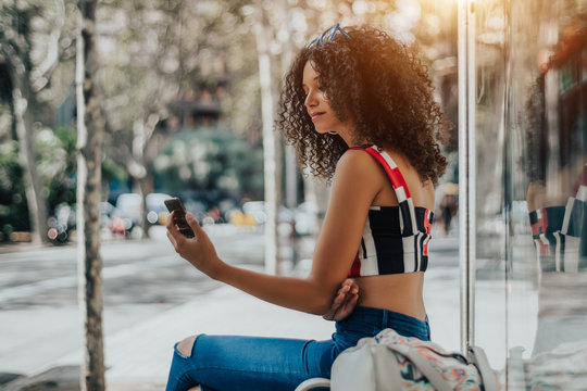 Charming African-American female is sitting on the bench inside of urban bus stop and having a video call via her smartphone; a dazzling woman with curly Afro hair with cellphone is waiting for a bus - Powered by Adobe
