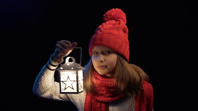 Little girl wearing knitted winter hat and scarf with lantern appearing in frame searching and waving head rejecting, over black background