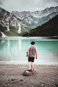 Mann mit hut und tank-top steht am ufer auf einem stein und blickt auf den plansee und die alpen