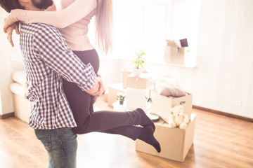Close up of young couple in a bright room. Young man is holding his wife on the hands. There are boxes with different stuff on the floor.