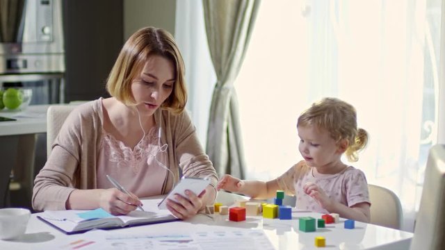 Medium shot of happy working mother talking on video call on mobile phone and writing in her planner while spending time with cute toddler girl playing with building blocks