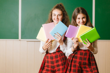 two funny schoolgirls in school uniform are standing with books on the background of the school board