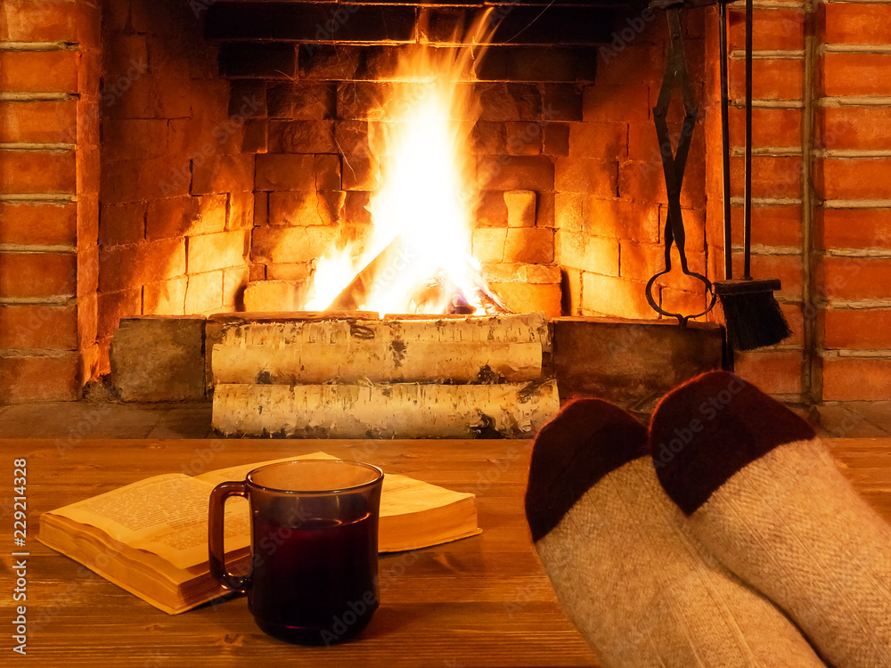 Wall mural cup of tea, book, women's feet in warm socks on a wooden table opposite a burning fireplace