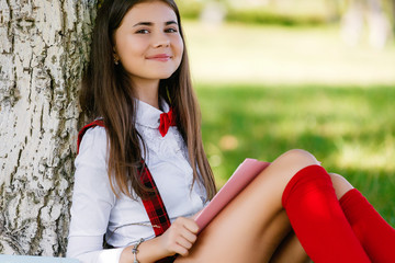 young schoolgirl in school uniform sitting on the grass under a tree with books
