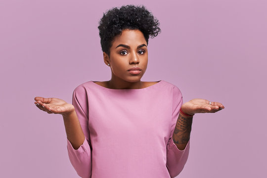 Uncertain African American female with dark skin, shruggs shoulders and asks something, has clueless expression, isolated on lavender background. Woman faces dilemma