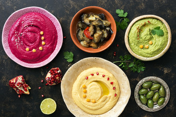 Colorful hummus in bowls on a dark rustic background, traditional hummus, hummus with basil, beet hummus. Top view, flat lay. Clean eating, dieting, vegetarian party food