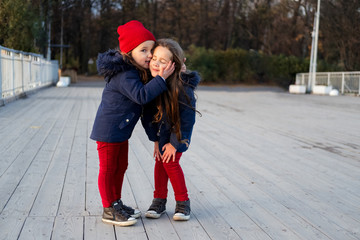 Two happy children hugging and kissing in autumn park. Close up sunny lifestyle fashion portrait of two beautiful caucasian girls outdoors, wearing cute trendy outfit. Sisters relationship