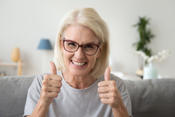 Excited mature woman in glasses sit on couch at home showing thumbs up satisfied with service,...