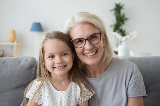Portrait of smiling cute little girl posing with grandma hugging and cuddling at home, happy grandmother and granddaughter spend time together look at camera making family picture