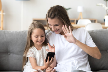 Obraz na płótnie Canvas Happy mom and daughter make video call on smartphone talking with dad or relatives, smiling little girl and mother waving at cellphone, having skype conversation from home. Kids and technology concept