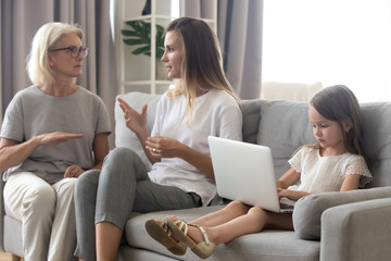 Small girl sit alone using laptop while mom and granny talk aside, having adult conversation, little daughter entertain herself watching video on computer, mother and grandmother chatting