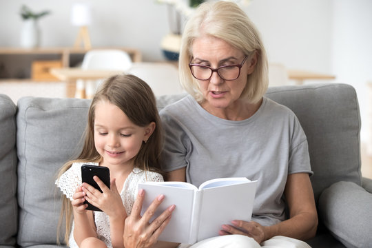 Grandmother read book to little granddaughter interest her in literature, distracted small girl addicted to smartphone play game or watch video on cell, not listening to granny. Generation gap concept