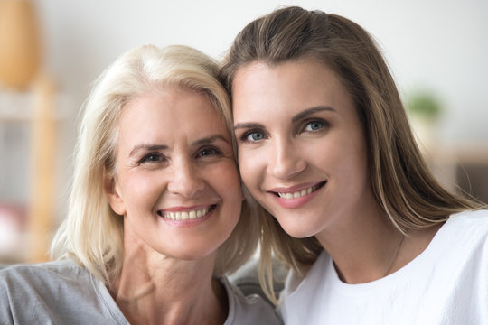 Portrait of smiling adult daughter and elderly mother look at camera posing for family picture, happy young woman and aged mom touch cheeks having close tender moment at home together