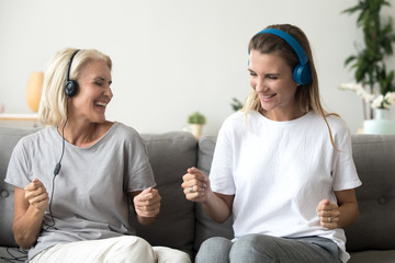Funny elderly mother and adult daughter listen to music in earphones having fun at home together,...