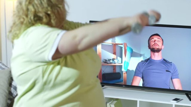 PAN of overweight woman in t-shirt holding light dumbbells and repeating exercise after online personal trainer on TV screen