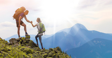 Young tourists with backpacks, athletic boy helps slim girl to clime rocky mountain top against bright summer sky and mountain range background. Tourism, traveling and healthy lifestyle concept.