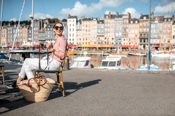 Young woman enjoying coffee sitting at the cafe outdoors near the harbour with beautiful buildings on the background in Honfleur old town, France - obrazy, fototapety, plakaty