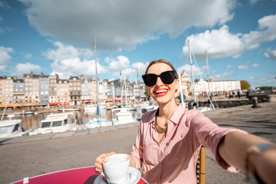 Young woman making selfie photo sitting at the cafe outdoors near the harbour in Honfleur old town, France