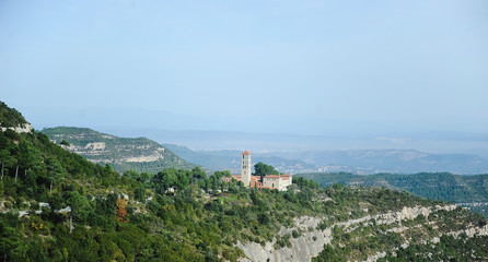 Fototapeta na wymiar Montserrat, Catalonia, Spain. Top View Of Hillside Cave Santa Cova De Montserrat Or Holy Cave Of Montserrat In Summer Day. Santa Maria De Montserrat Abbey.