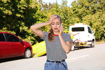 Woman talking on phone near broken car and tow truck outdoors