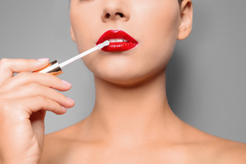 Beautiful woman applying red gloss on lips against gray background, closeup