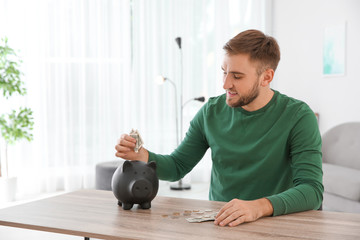 Young man putting coin into piggy bank at table indoors. Space for text