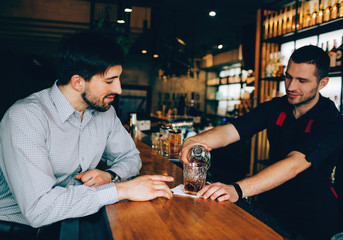 Customer sitting at the barman's stand and looking how barmen is pouring in some alcohol to the glass. He is controlling that process.