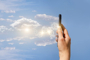 A hand holding a mobile phone emitting the mechanical details at the cloudy shiny background.