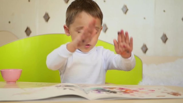 A naughty boy takes paint and claps his palms on the paper. Development, education, psychology, emotions of a child.