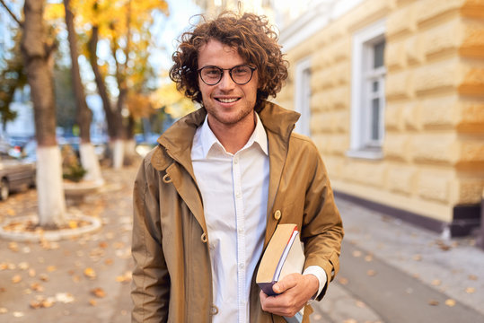 Portrait of handsome young man with books outdoors. College male student carrying books in college campus in autumn street background. Smiling guy with glasses and curly hair posing with books outside