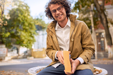 Happy handsome young man reading and posing with book outdoors. College male student carrying books in campus in autumn street. Smiling smart guy wears spectacles and curly hair reading books outside
