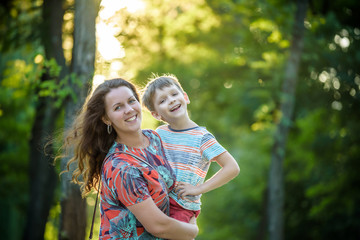 Fototapeta premium Beautiful woman and her cute little son are hugging and smiling, on nature park background. Boy is looking at camera. Concept of friendship between children and parents, happy family