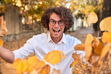 Horizontal autumn shot of happy handsome winner young man with glasses playing with leaves...
