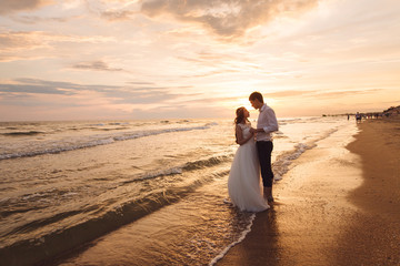 A beautiful couple of newlyweds, the bride and groom walking on the beach. Gorgeous sunset and sky. Wedding dresses, a white luxury dress for a girl. Family concept, honeymoon.