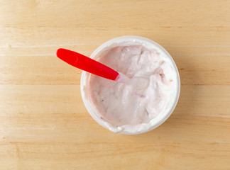 Top view of a container of strawberry rhubarb yogurt sweetened with honey plus a spoon in the food on a wood table.