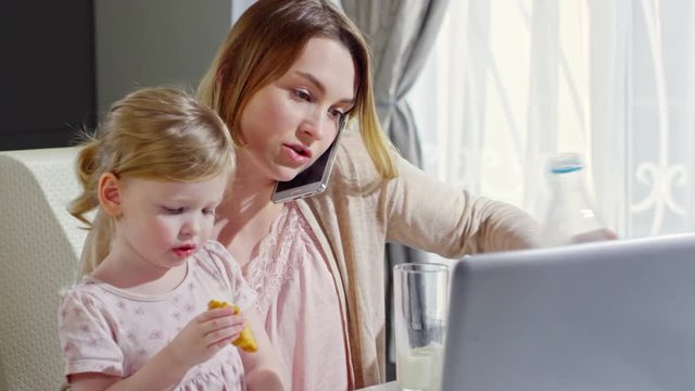 PAN of beautiful young mother talking on phone and pouring milk into glass and offering it to cute toddler girl sitting on her laps and eating croissant while watching cartoons on laptop