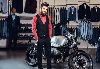 Obraz na płótnie Canvas Elegantly dressed man posing with hands in pockets near retro sports motorbike at the men's clothing store.