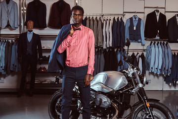 Fototapeta na wymiar African American young man dressed in elegant formal wear posing near retro sports motorbike at the men's clothing store.