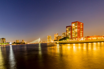 Fototapeta premium Night cityscape of buildings, yellow lights reflected in the water and the Erasmus cable-stayed bridge over the Maas river in the background in Rotterdam, clear sky in the Netherlands